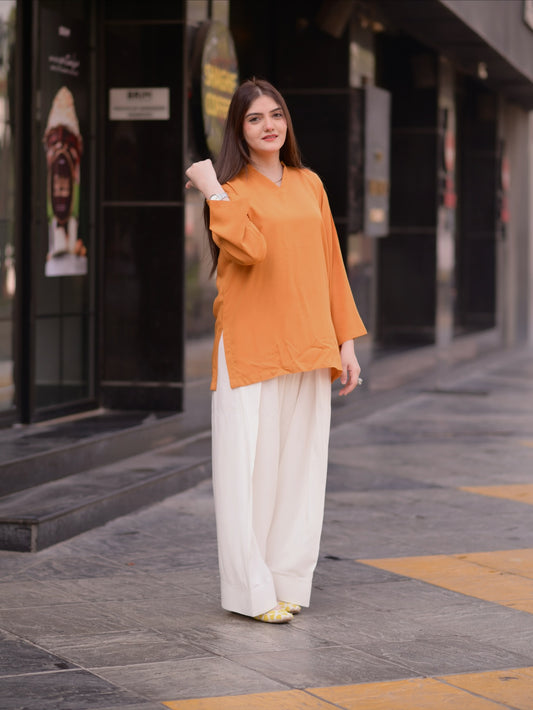 A woman stands on a city sidewalk wearing a loose mustard-orange long-sleeve tunic top with side slits and wide-leg off-white trousers.