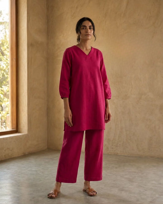 A woman modeling a bright magenta two-piece linen kurta and trouser set in a rustic room with textured beige walls and a window.