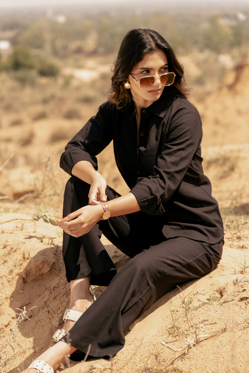 Woman in black outfit and sunglasses sitting on a rocky surface with a desert landscape in the background