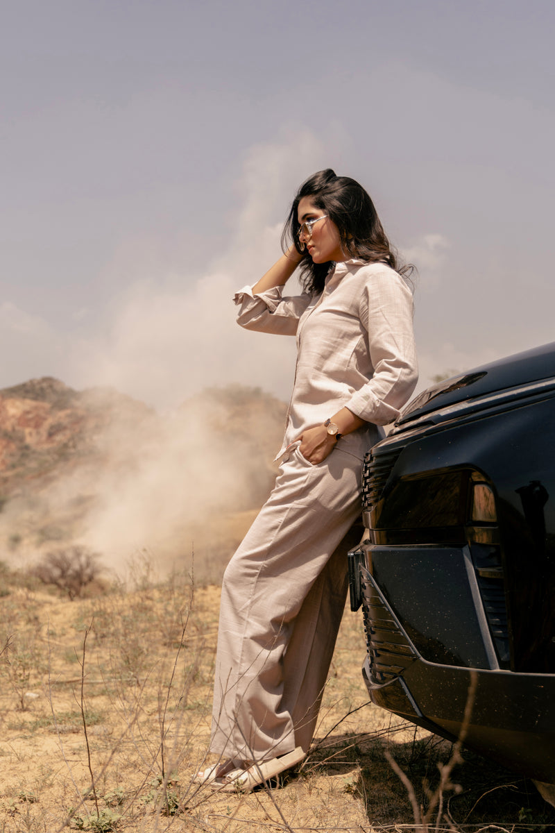 Woman in a beige outfit standing next to a car in a desert-like setting
