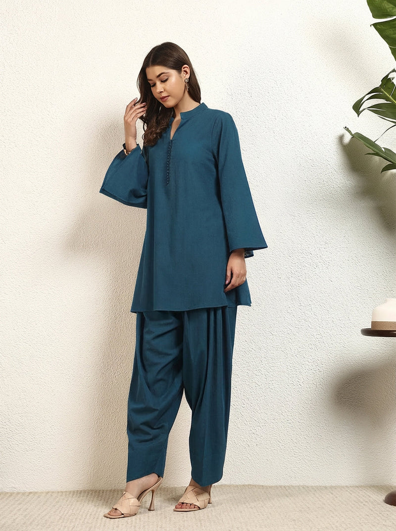 Woman wearing a teal outfit standing against a white wall.
