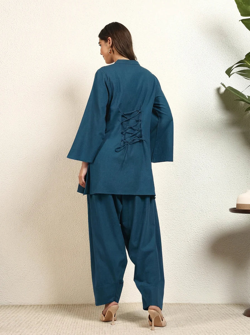 Woman wearing a teal outfit with a lace-up back, standing against a white wall.