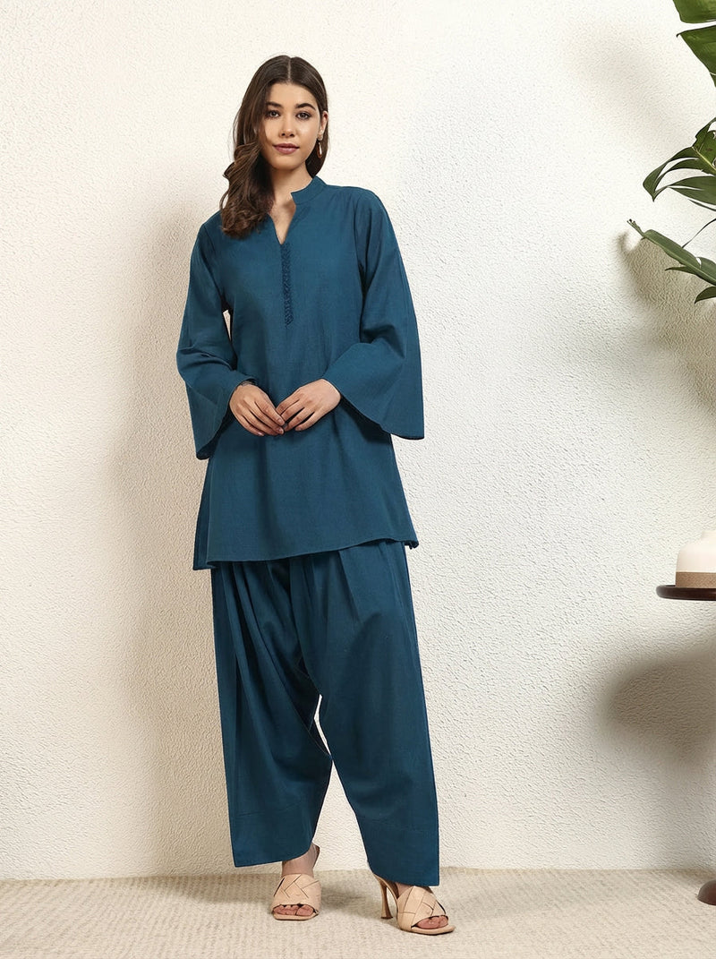 Woman wearing a teal outfit standing against a white wall with a plant in the corner.