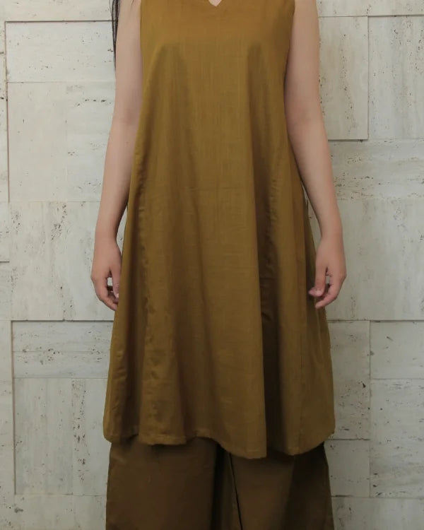 Person wearing a mustard yellow sleeveless top and brown pants against a light stone wall.
