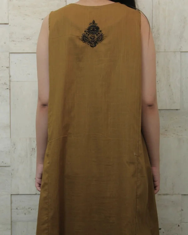 Brown sleeveless dress with black embroidery on the back against a light stone wall.