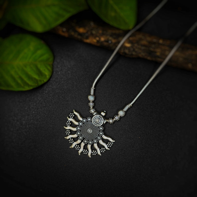 Silver sun-shaped pendant necklace on a dark surface with green leaves in the background
