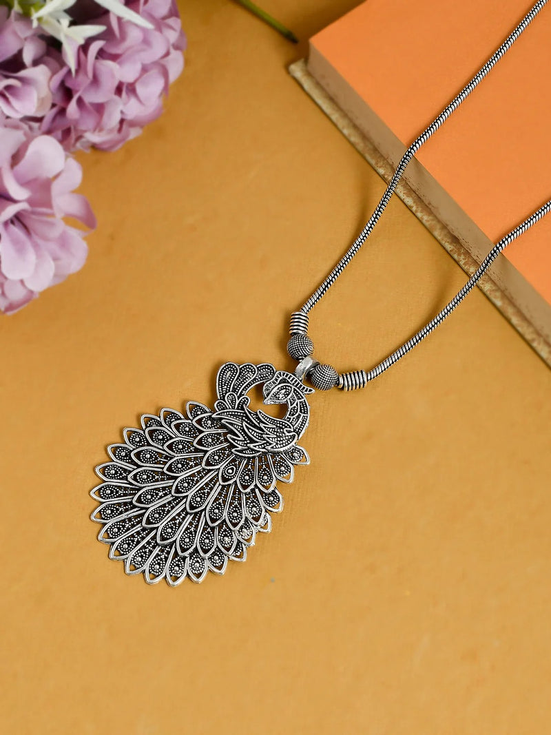 Silver peacock feather pendant necklace on a yellow background with pink flowers.