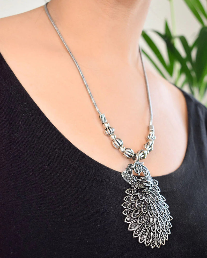 Silver necklace with a detailed pendant on a black top against a blurred green plant background