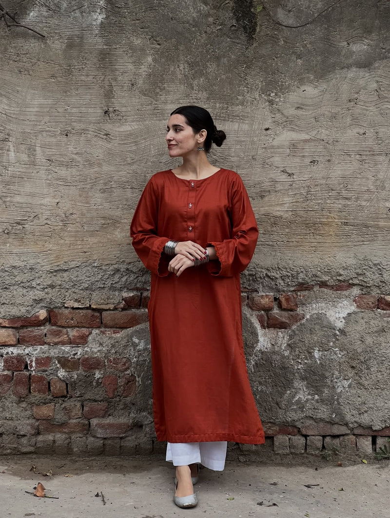 Woman in a rust-colored kurta standing against a textured wall.