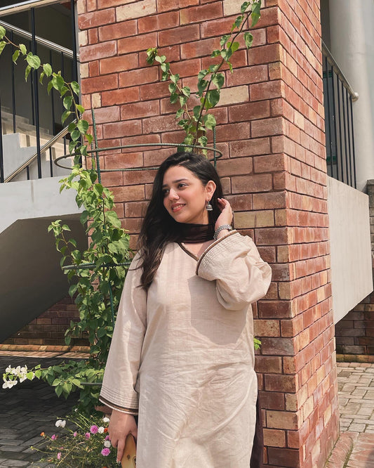 Woman in a beige outfit standing against a brick wall with greenery