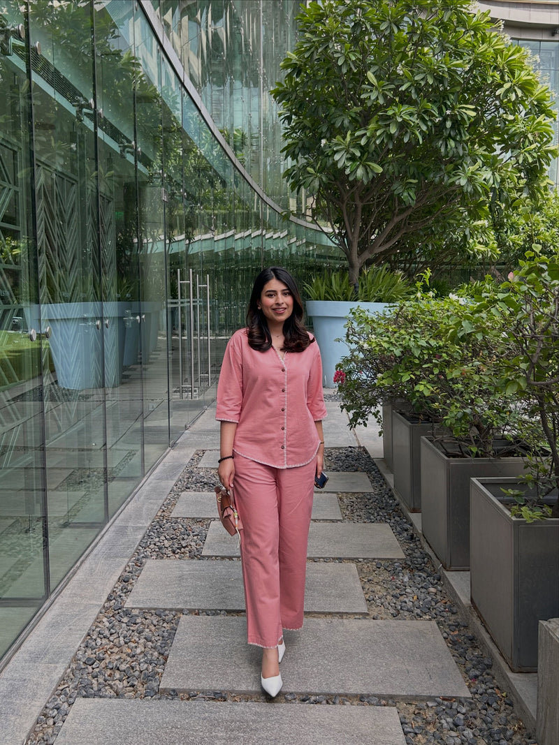 A woman wearing a matching dusty pink two-piece set consisting of a button-up shirt and wide-leg trousers, walking along a stone path in a modern garden corridor with glass walls and potted plants.