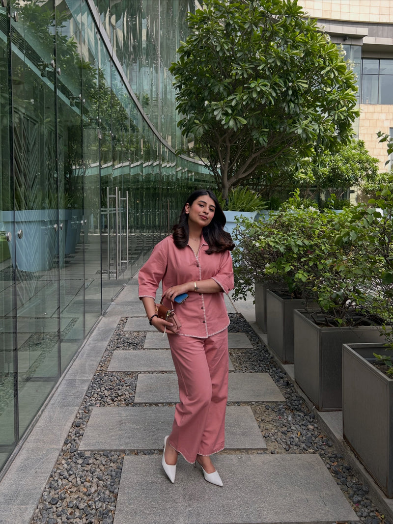 A woman wearing a matching dusty pink pajama set with a button-up shirt and wide-leg pants, standing on a stone path in a modern outdoor corridor with glass walls and potted plants.