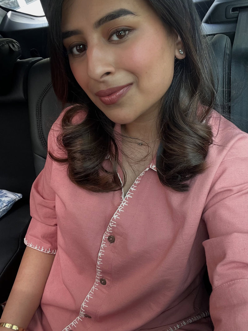 A woman wearing a dusty pink short-sleeve top with white embroidered trim and brown buttons, sitting in a black car interior.