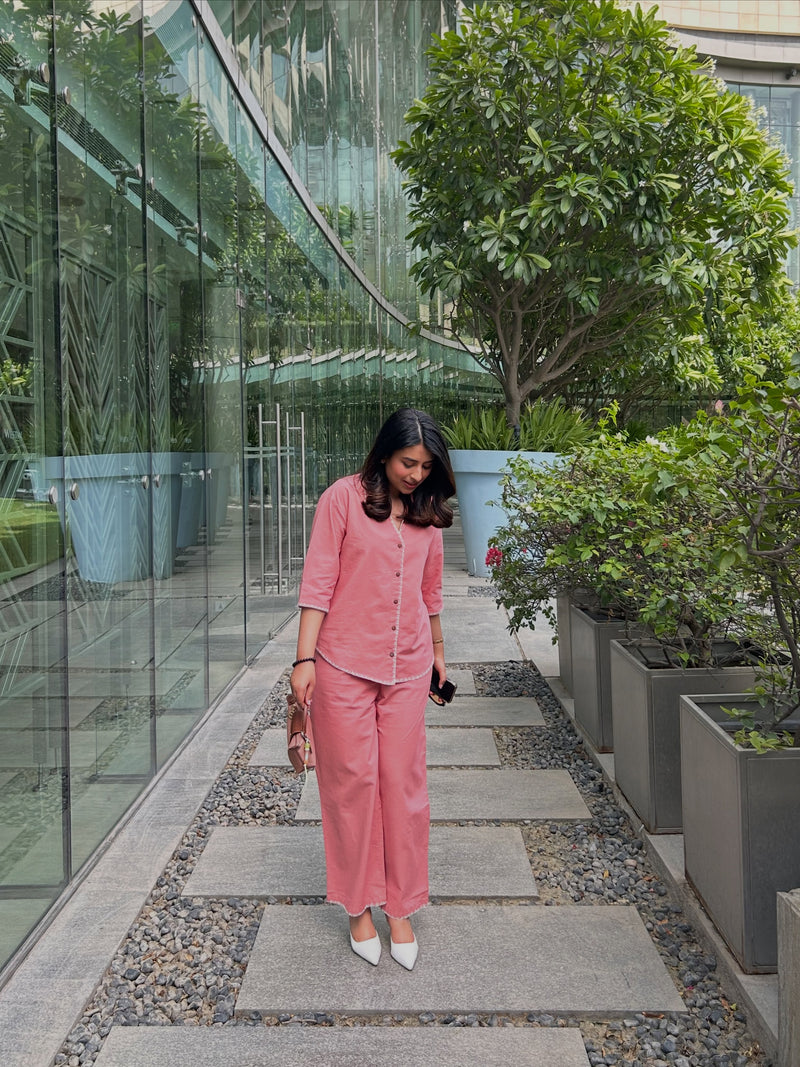 A woman wearing a matching pink two-piece button-up shirt and wide-leg trousers walks along a stone path bordered by green plants and a glass wall.