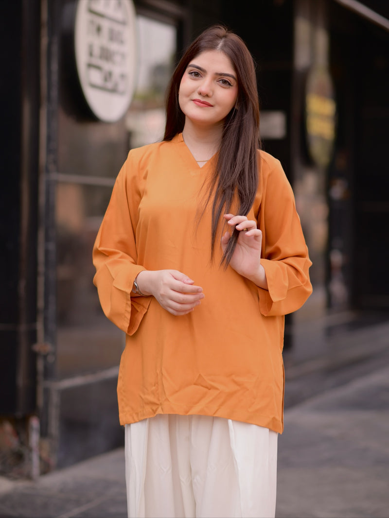 A woman wearing a loose-fitting orange tunic top with wide sleeves and white pants, standing outdoors in front of a blurred storefront background.