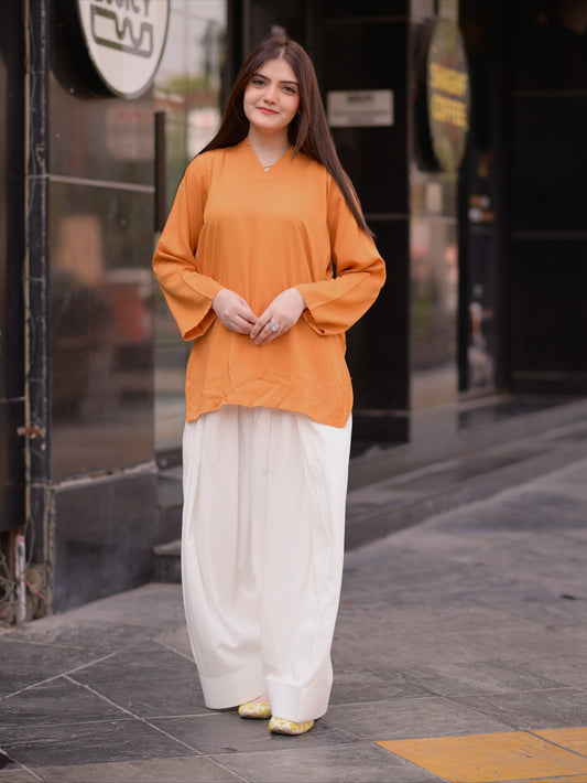 A woman standing on a city sidewalk wearing a loose mustard-orange long-sleeve top and wide-leg white pants with gold shoes.