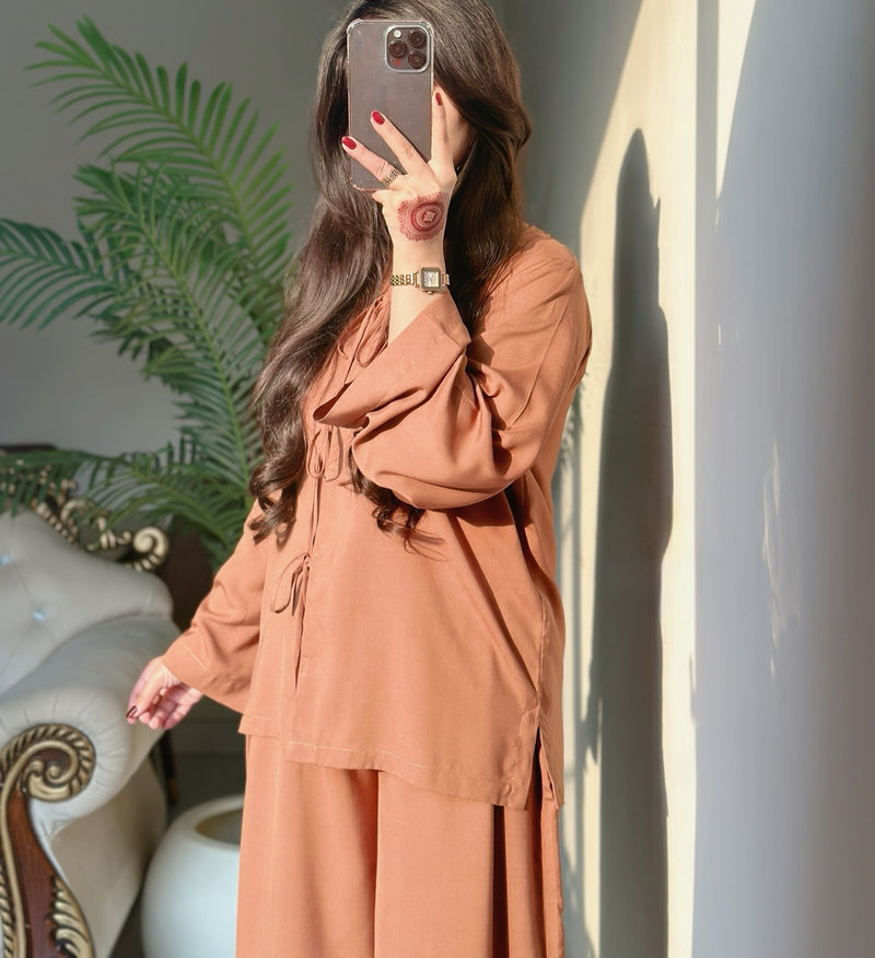 Person wearing a brown outfit taking a mirror selfie with a plant and decorative elements in the background.