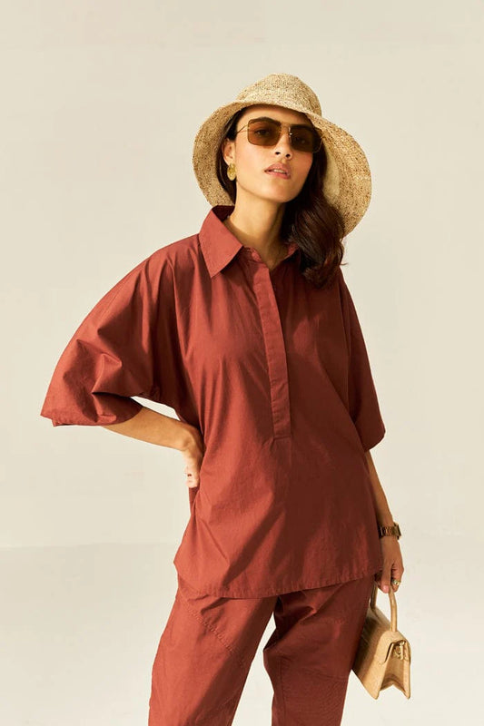 A woman modeling a matching rust-red short-sleeve button-up shirt and trousers set, styled with a wide-brim straw hat, sunglasses, and a small beige handbag against a plain light background.
