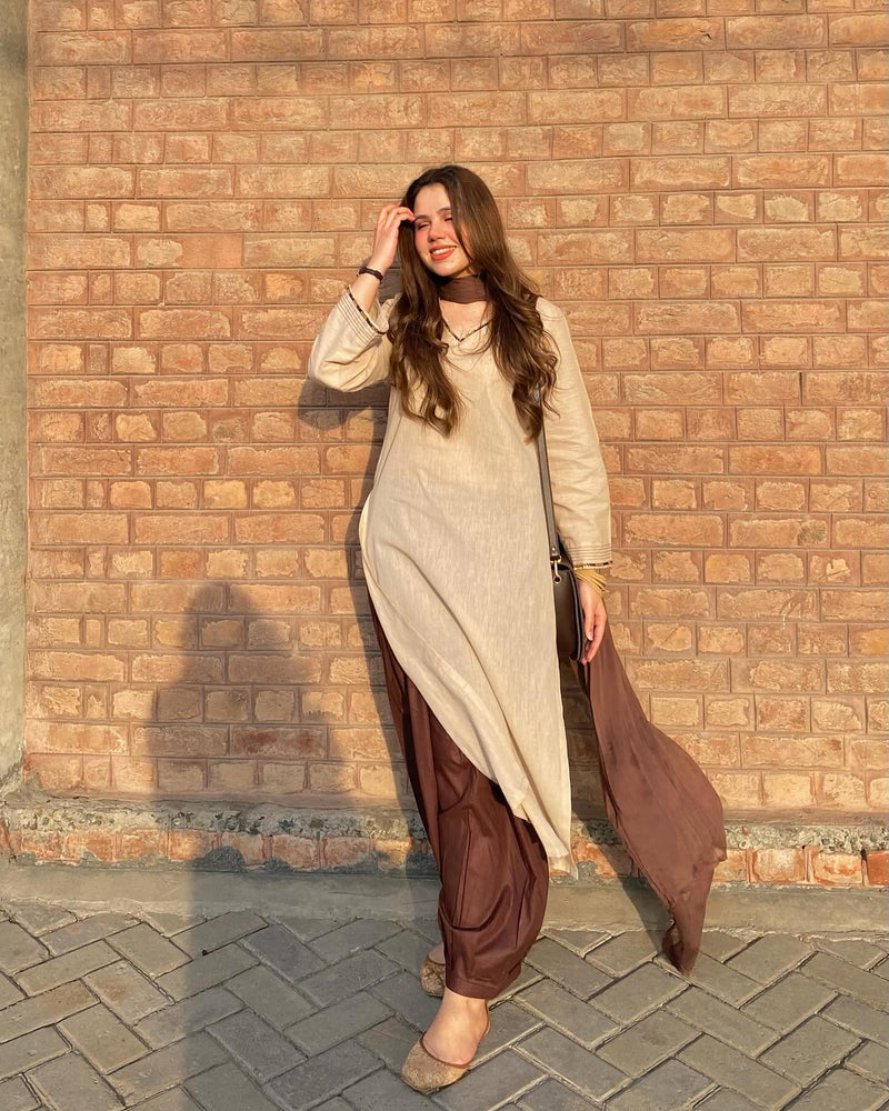 Woman in a beige top and brown pants standing against a brick wall.